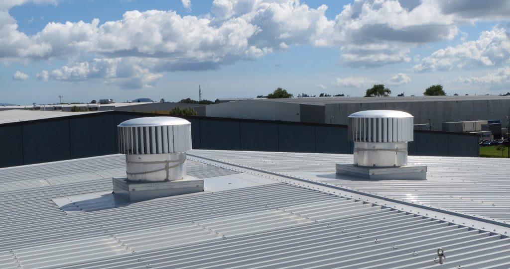 Two silver-colored turbine roof vents on a corrugated metal roof under a partly cloudy sky, with industrial buildings visible in the background.