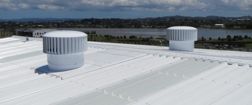 A white industrial building with a corrugated metal roof, featuring two large, white roof ventilators, with a body of water and distant hills under a cloudy sky in the background.